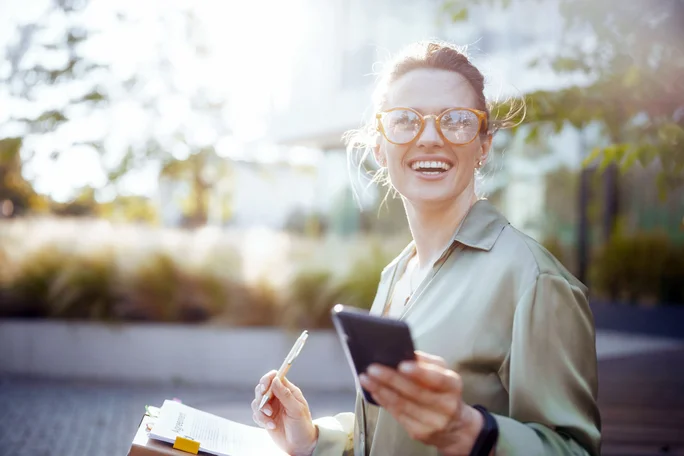 Imaginea prezintă o persoană aflată în aer liber, ținând un smartphone într-o mână și un clipboard cu un pix în cealaltă. În fundal se observă vegetație și o clădire aflată în plan secund.